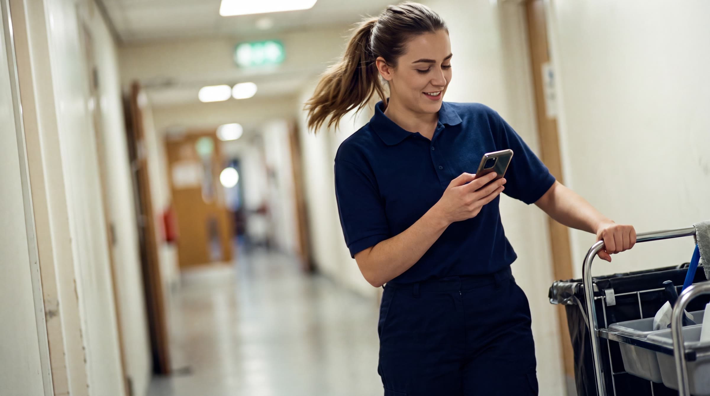A young supervisor in uniform smiles at her phone while pushing the cleaning trolley along a corridor. Illustrative photo.